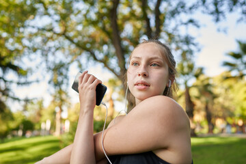 a beautiful red-haired girl with green eyes in a medium shot stretches her muscles to be able to start playing a sport in a park in the center of the city