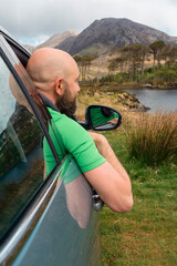 Bald male driver looking at Twelve pines island from car window, county Galway, Connemara region, Ireland. Outdoor activity and travel concept. Mountains in the background. Blue sky