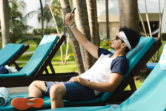 Young hispanic man sitting in a long chair by the pool taking a picture - young man on vacation taking a selfie while spending time relaxing