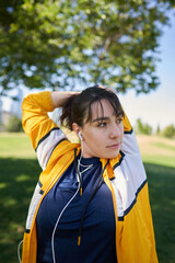 A beautiful white woman with brown hair and a sports suit with a yellow jacket prepares for exercise and exercises her muscles to avoid cramps in a park near the city wearing a Covid 19 mask     