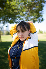 A beautiful white woman with brown hair and a sports suit with a yellow jacket prepares for exercise and exercises her muscles to avoid cramps in a park near the city wearing a Covid 19 mask     
