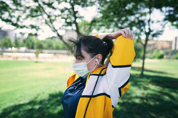 A beautiful white woman with brown hair and a sports suit with a yellow jacket prepares for exercise and exercises her muscles to avoid cramps in a park near the city wearing a Covid 19 mask     