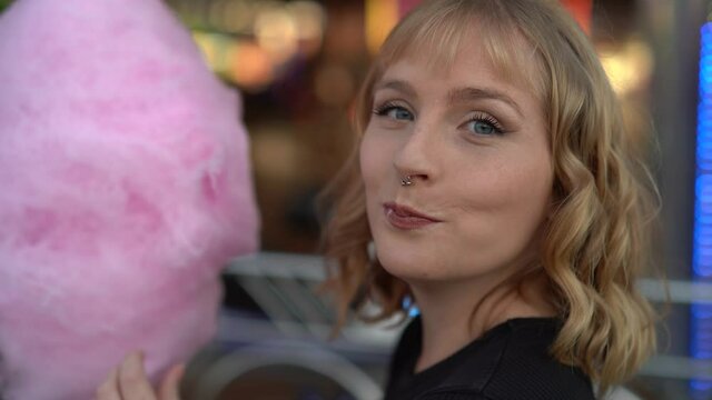 Primer plano de chica rubia guapa con ojos azules comiendo algod&oacute;n de az&uacute;car mientras sonr&iacute;e en zona de atracciones de una feria