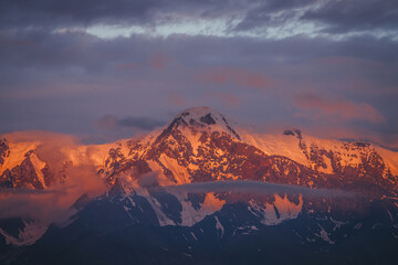 Scenic mountain landscape with great snowy mountains lit by dawn sun among low clouds. Awesome alpine scenery with high mountain pinnacle at sunset or at sunrise. Big glacier on top in orange light.