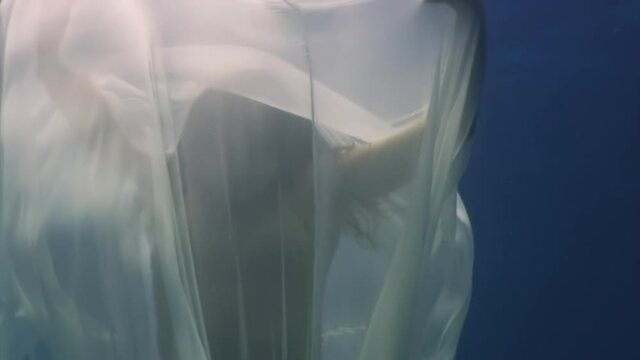 Young woman underwater model in white cloth on background of blue water. Concept of creativity and art of creating images.