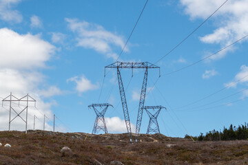 Large transmission lines and metal towers moving electricity, conductors, and distribution wires for high voltage electricity. The poles have a blue sky in the background with white clouds.