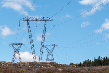 Large transmission lines and metal towers moving electricity, conductors, and distribution wires for high voltage electricity. The poles have a blue sky in the background with white clouds.