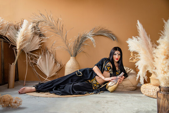 a beautiful young woman with black hair dressed in the costume of scheherazade an oriental beauty lies on the floor with a jug