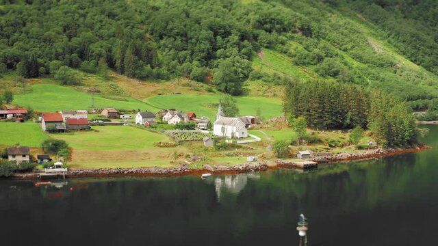 Aerial view of the village of in Bakka, Norway. A small village sitting on the coast. Green farm fields and steep, forest-covered mountains. Small tidy houses and a white wooden church.