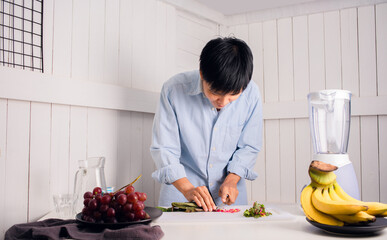 Enjoying Asian man slicing vegetables preparing to Make a Smoothie with Swiss Chard, banana, and red grape at a white kitchen home. people are taking care of themselves.