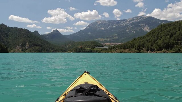 First person view looking from yellow kayak swaying on waves of emerald lake. Mountain and forest landscape on summer day