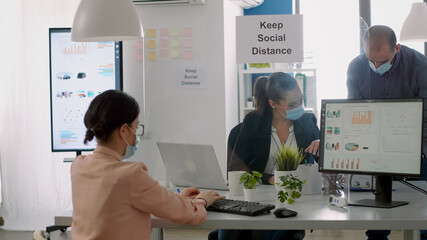 Group of business people with face masks working in new normal company office analysing financial project during coronavirus global pandemic. Coworkers keeping social distancing to avoid virus disease