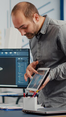 Industrial designer holding tablet discussing with woman engineer while working in CAD program, designing new components, comparing 3D prototype with project from notepad, using pc with two monitors