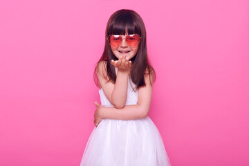 Little girl in a smart white dress and heart-shaped glasses looks at camera, smiles and sends an air kiss, expressing positive emotions, isolated over pink background.