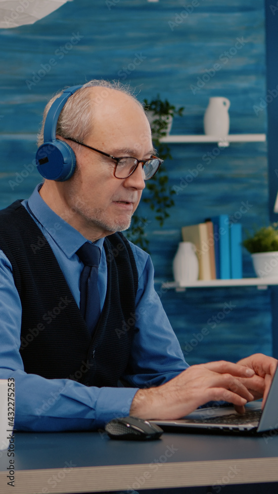 Poster focused old entrepreneur with headphones listening music while checking graphics writing on laptop w