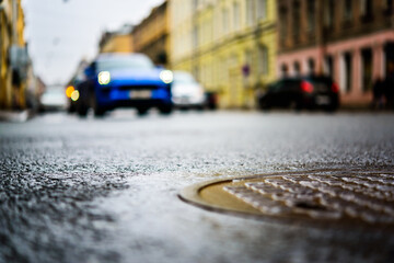 Rainy day in the big city, the headlights of the approaching blue car on the road. Close up view of a hatch at the level of the asphalt