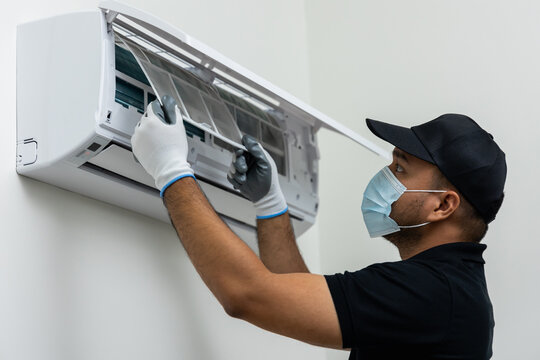 Air Conditioner Service Indoors. Air Conditioner Cleaning Technician He Opened The Front Cover And Took Out The Filters And Washed It. He In Uniform Wearing Rubber And Mask.