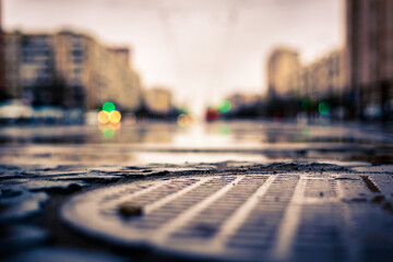 Rainy day in the big city, an empty road with glowing lights from cars. Close up view of a hatch at the level of the asphalt
