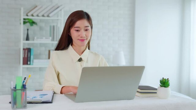 Asian young adult businesswoman do remote stay work from home sit at desk smile enjoy telework activity. Spend time chatting in social network use laptop working at home. New normal lifestyle concept.