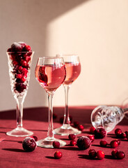 red berries scattering from a crystal glass lying on the table among glasses with pink alcoholic drink, light and shadow, cranberry's or cherry's vodka, liqueur
