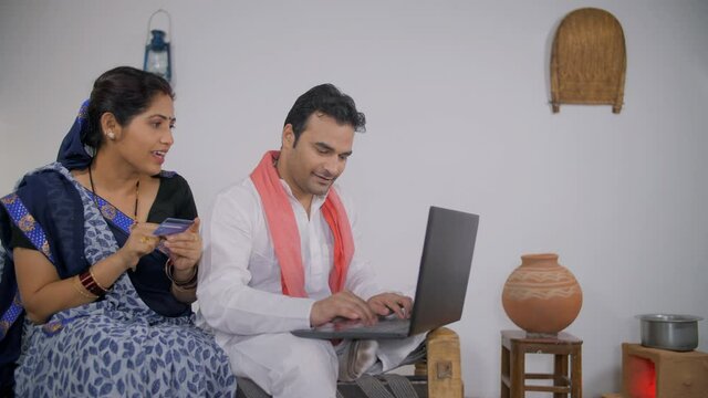 Indian village couple doing card payment using a laptop - online shopping concept. Young beautiful wife holding a debit/credit card in her hand and telling her husband card details to do online pay...