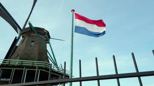 Dutch red, white and blue flag waving in the wind with an old dutch windmill and blue sky in the background during a sunny day. Slow-motion clip.