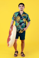 Young Asian man in colorful printed Hawaiian shirt smiles with happiness while holding swim ring. Full body studio portrait on yellow background. Summer holiday travel concept.