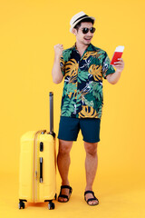 Enjoyed young Asian man in colorful Hawaiian shirt holds passport and make a winner gesture near suitcase. Full body studio portrait on yellow background. Happy summer vacation travel concept.