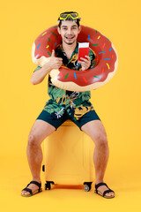 Excited young Asian man in colorful Hawaiian shirt holds passport and swim ring while sitting on yellow suitcase. Full body funny studio portrait on yellow background. Summer holiday travel concept.