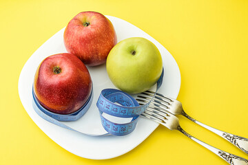 Red and yellow apples, wrapped with a centimeter tape on a white plate, on a yellow background, next to two table forks. Healthy food, calorie content. Selective focus. Top and side view.