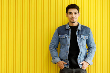 Trendy attractive Asian man in denim jacket standing against yellow background. Medium shot portrait, fashionable urban concept. © Bangkok Click Studio