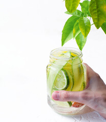 A woman's hand holds glass mug with handle with refreshing detox lemonade with sliced lime, lemon, cucumbers and red orange on white table. 