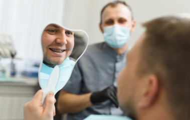 Installation of braces. The male patient looks in the mirror at the new braces for bite correction and smiles. Satisfied patient of dentistry. Male doctor in the background