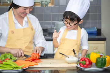 Asian woman young mother with son boy cooking salad food with vegetable holding tomatoes and carrots, bell peppers on plate for happy family cook food enjoyment lifestyle kitchen in home