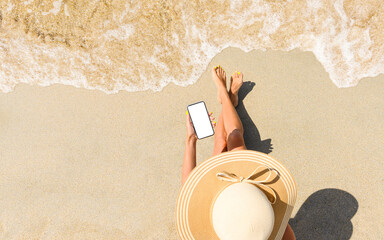 A girl in a straw hat sits on the beach with a phone in her hand. View from above. Phone with blank...