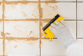 the hands of the master apply a brown grout with a spatula on the wall lined with white tiles, finishing work in the room