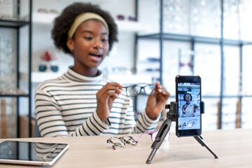 African blogger influencer recording video blog speaking and looking at smartphone on tripod at optical store, recording and share on social media live streaming Internet, selective focus