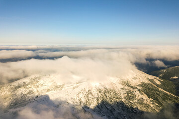 Aerial view from above of white puffy clouds covering snowy mountain tops in bright sunny day.