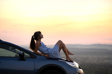 Happy young woman driver in blue dress laying on her car hood enjoying warm summer day. Travelling and vacation concept.