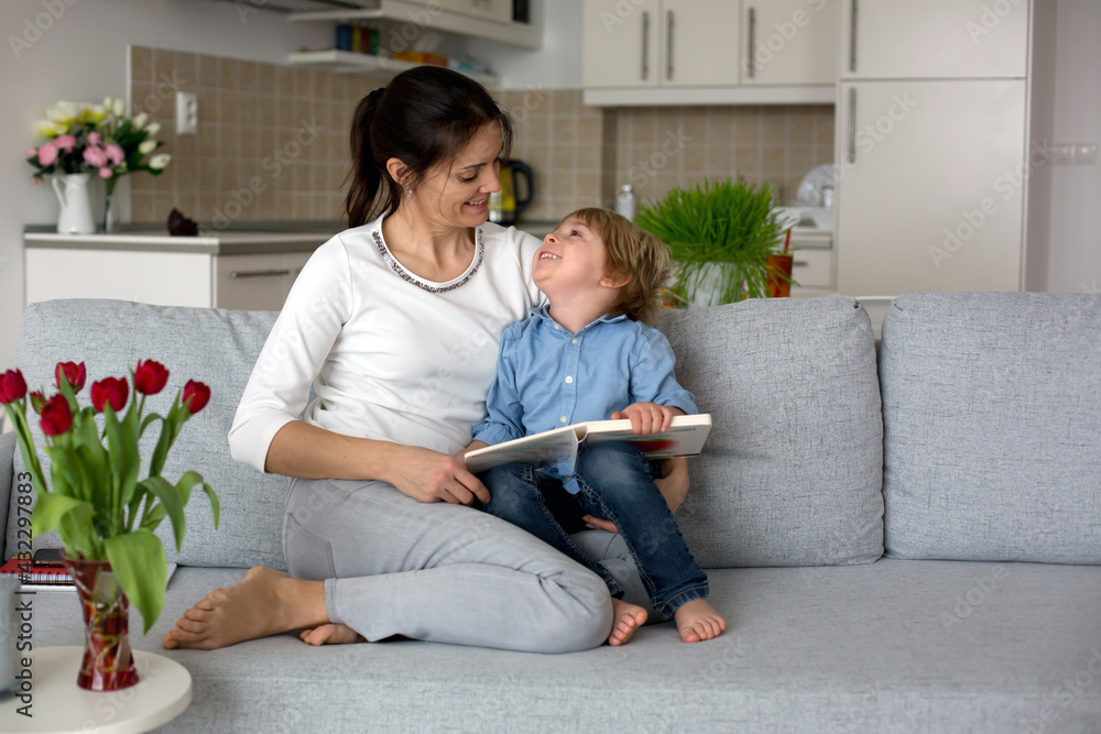 Wall mural Sweet boy and his mom, reading a book at home, sitting on the couch, relaxing, mom working from home