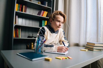 Low-angle view of thinking pupil boy thoughtful writing in notebook with pen sitting at desk near window. Close-up of child schoolboy doing homework at home.