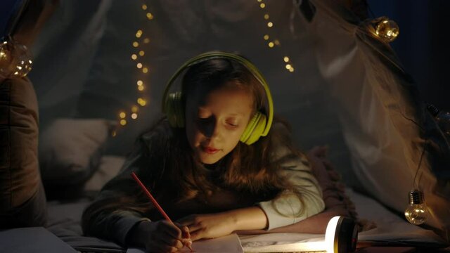 Young girl in headphones drawing with colored pencils while lying on floor in cosy decorative tent at home. Teen singing and listening to music while spending free time in evening.