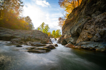 Mountain river with small waterfall with clear turquoise water falling down between wet boulders with thick white foam on autumn bright day.