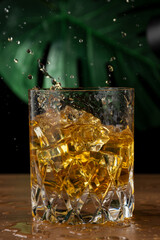 Close-up of glass with whiskey and ice splashing, on wooden table, black background with illuminated green leaf, vertical