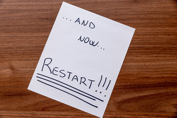 White sheet with the words "now ... restart!" with black ink, placed on wooden surface. 
