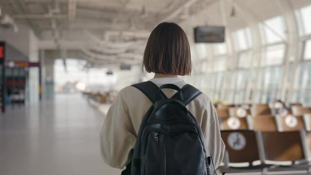 Back view of the short haired young woman walking through the empty airport with her backpack while waiting for the arrival of the plane