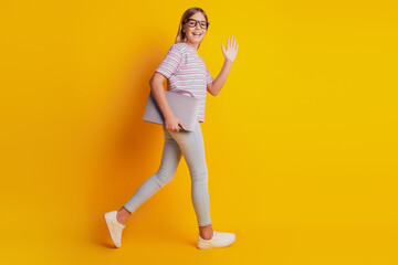 Fototapeta na wymiar Photo of young kid girl go hold laptop wave hand isolated over yellow background