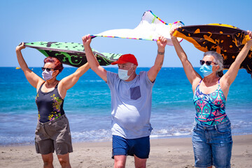 Three senior people enjoying the beach together holding towels in the wind, wearing face mask due to coronavirus - active retired seniors and vacation concept