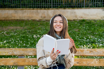 Young woman in white electronic headphones smiling and listening online audiobook in nature. Enjoying music or leisure podcast. Distance education. Storytelling. Text space