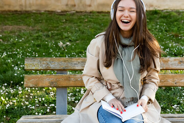 Young woman in white electronic headphones laughing and listening online audiobook in nature....
